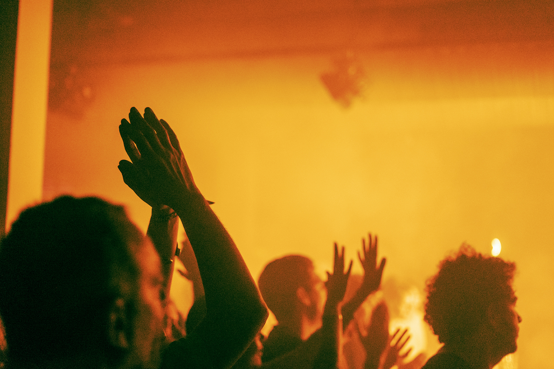 An orange, yellow photo of Le Guess Who audience. Some of the audience enthusiastically raise their hands.